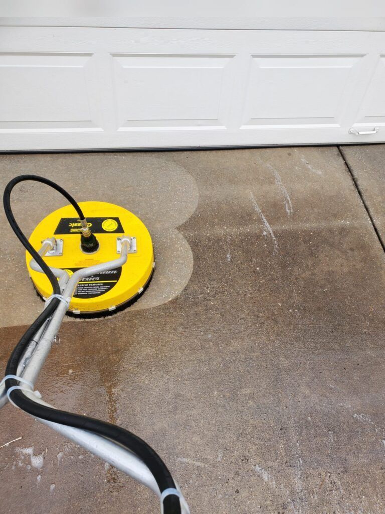 A yellow circular surface cleaner is being used to deep clean a dirty concrete driveway in front of a white garage door.