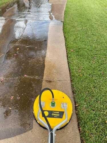A yellow surface cleaner tool operates on a concrete driveway, leaving a clean path compared to the adjacent dirty surface.