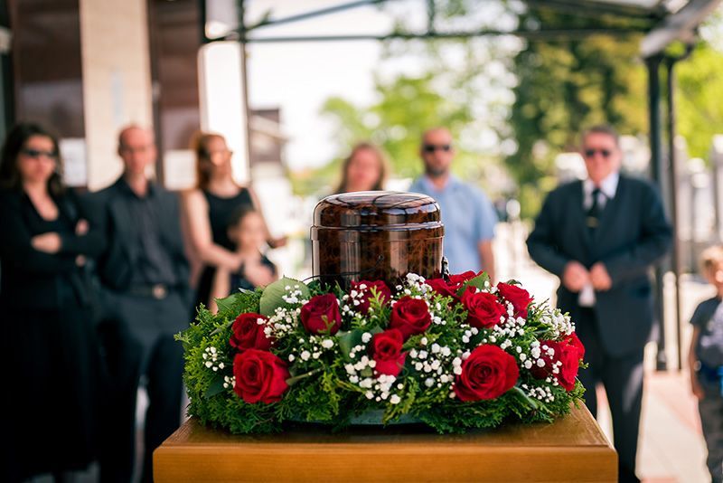 A dark urn sits on a wooden pedestal surrounded by red roses and greenery, with a group of blurred mourners in the background.