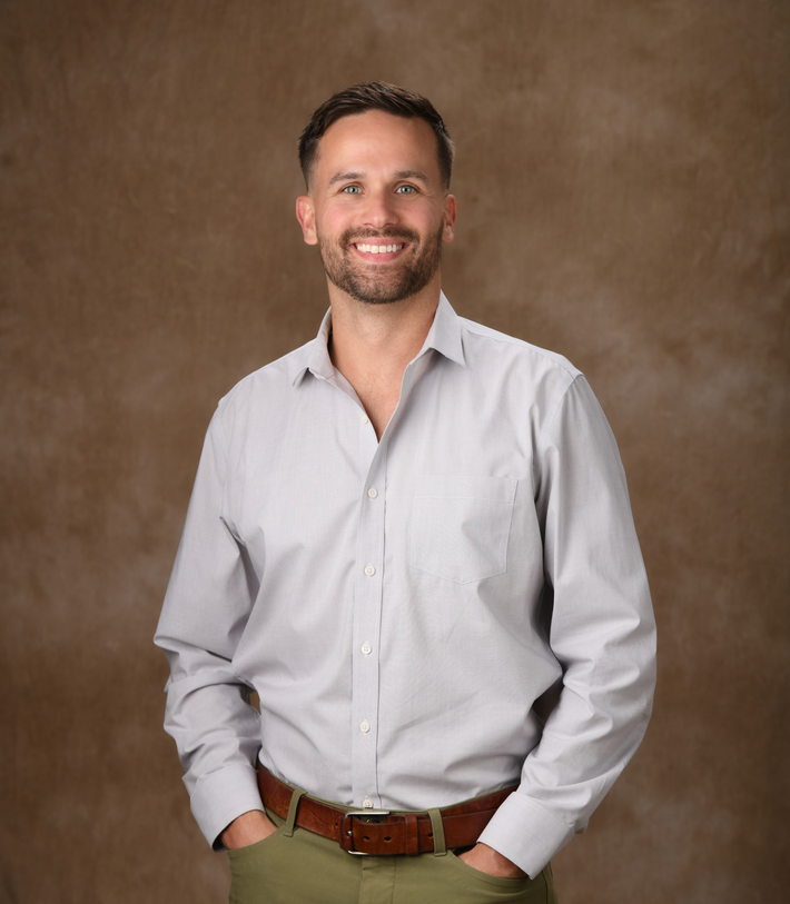 Smiling man in a light gray shirt and khaki pants, standing against a brown studio backdrop.