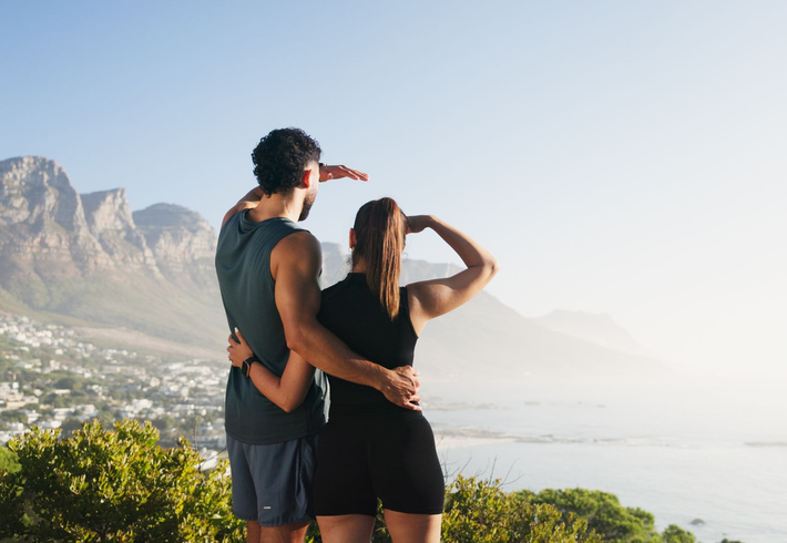 Two people standing on a coastal overlook, hugging and looking out over the ocean at sunrise.