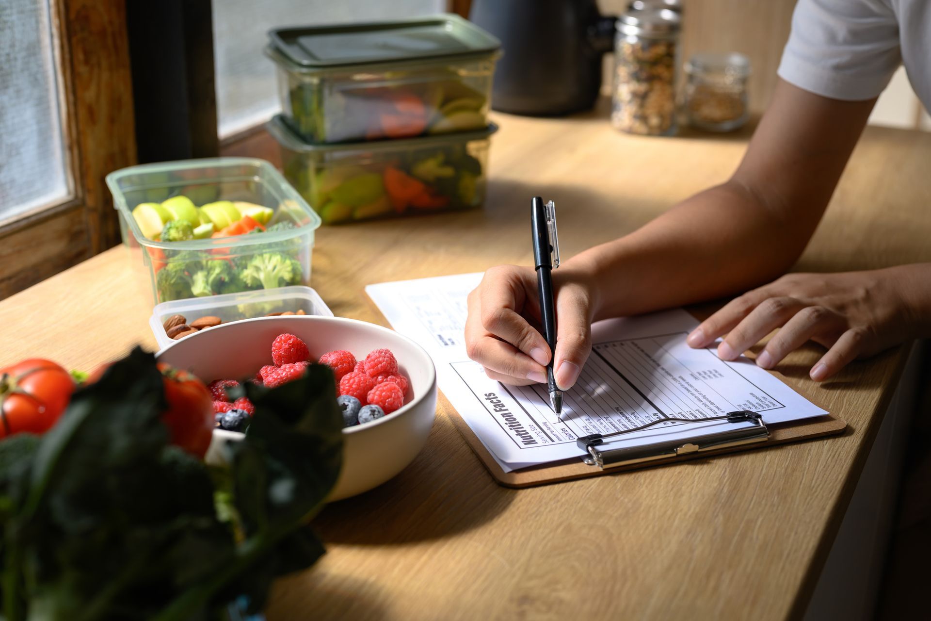 Person writing in a notebook beside fresh produce and meal-prep containers on a wooden table