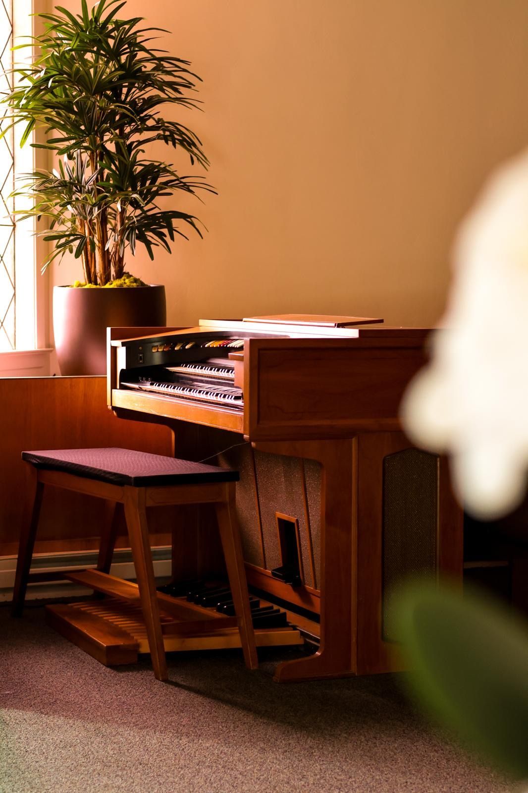 A small wooden piano beside a window, with a bench and potted plant in a warm-lit room.