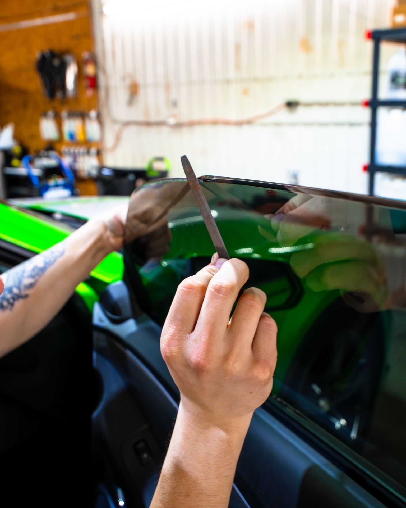 Hands applying window tint to a green car in a garage
