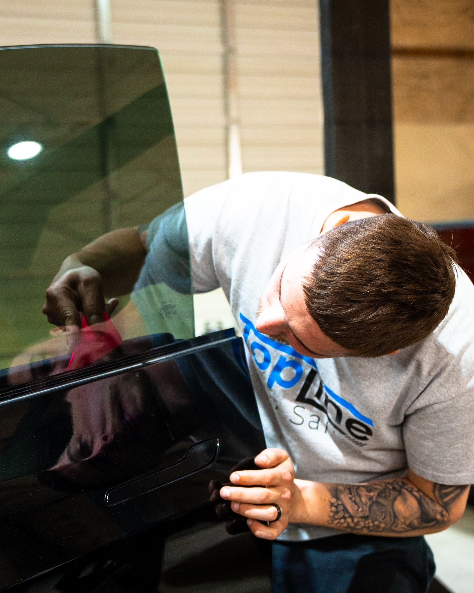 Person installing a tinted glass panel, wearing a white shirt with blue text and visible arm tattoos.