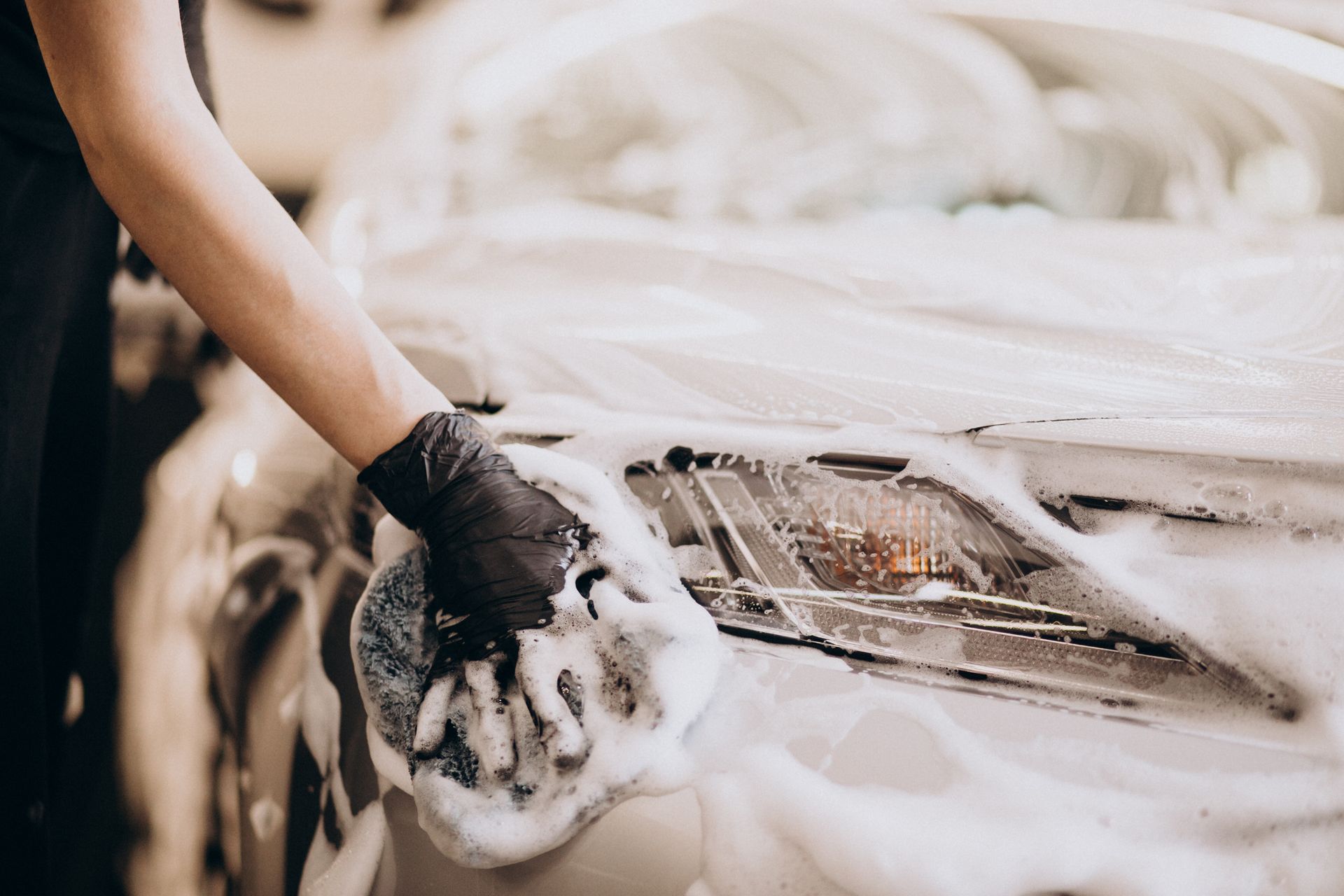 Hand wearing a black glove washing a white car’s headlight with soapy foam