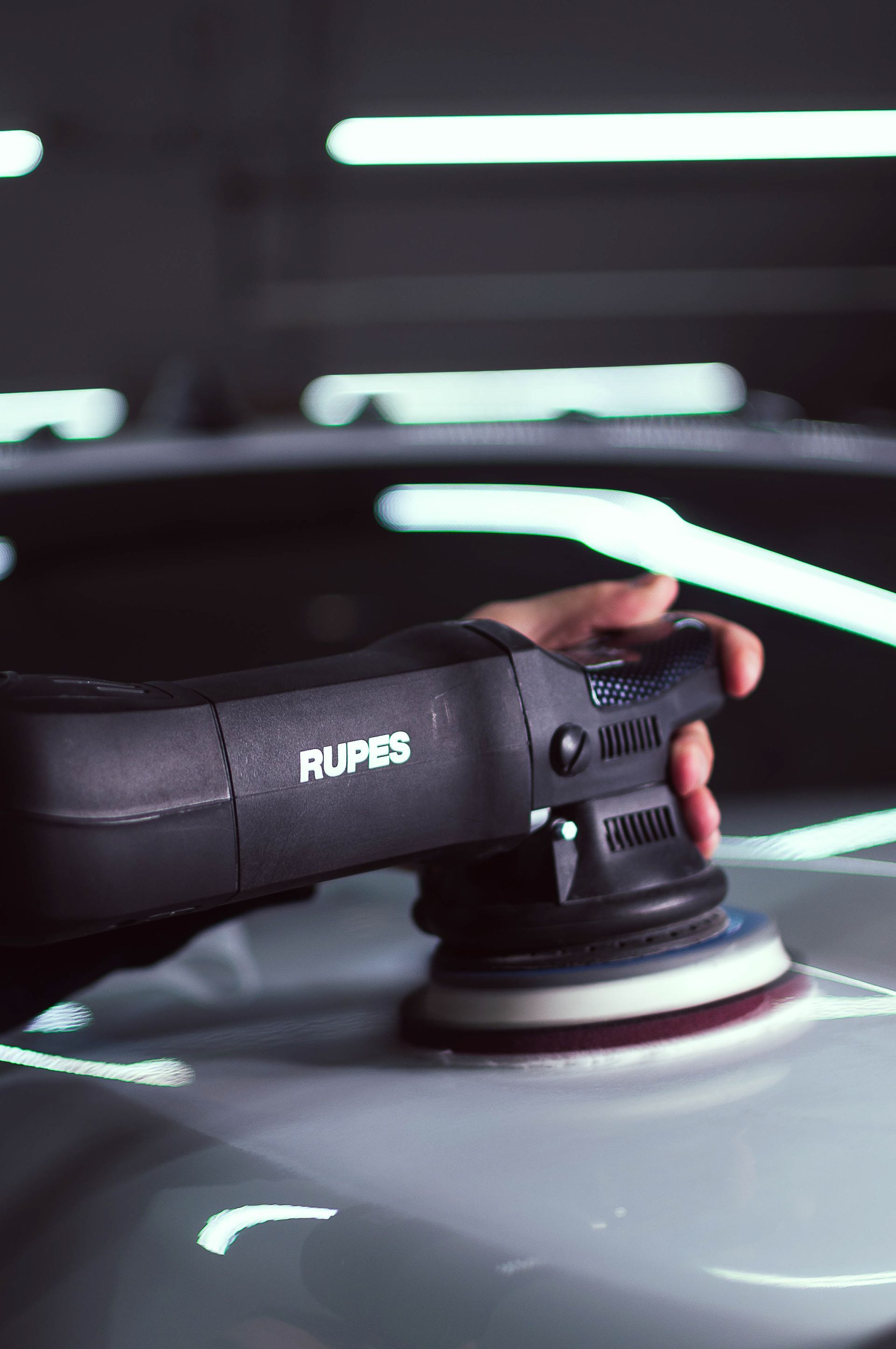 Hand polishing a gray car with a Rupes electric buffer in a dark garage