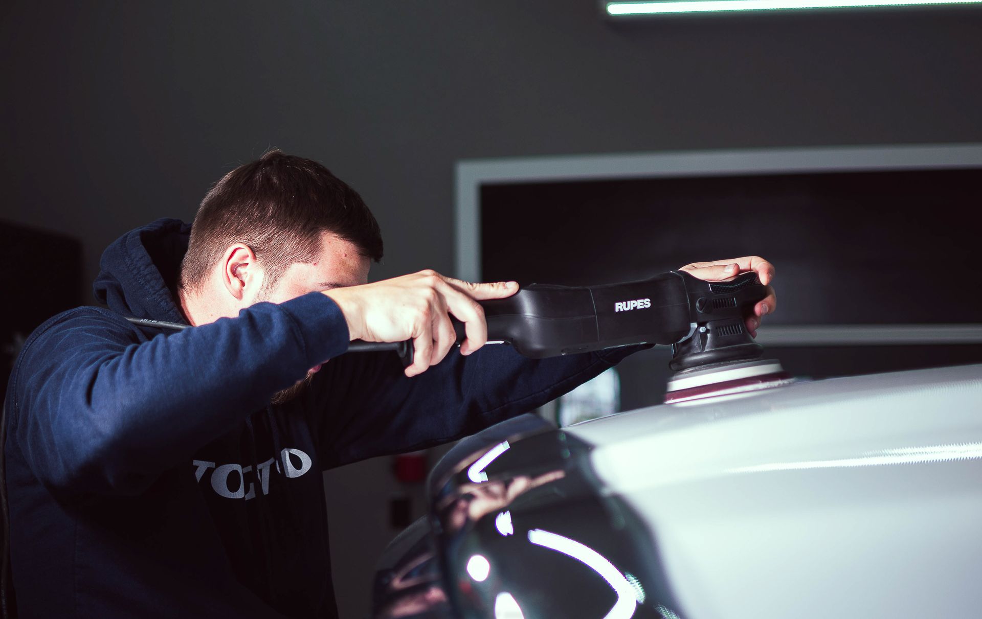 Person polishing a car with a cloth in a dim garage, close-up on the vehicle’s hood.