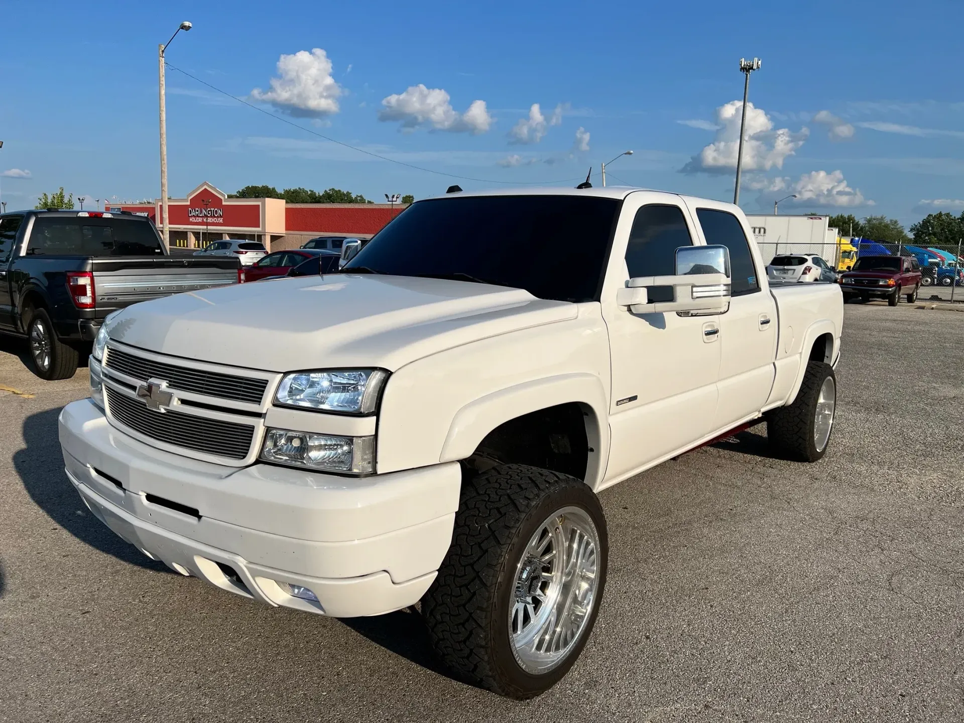 White lifted pickup truck parked on a gravel lot under a blue sky with other vehicles in the background