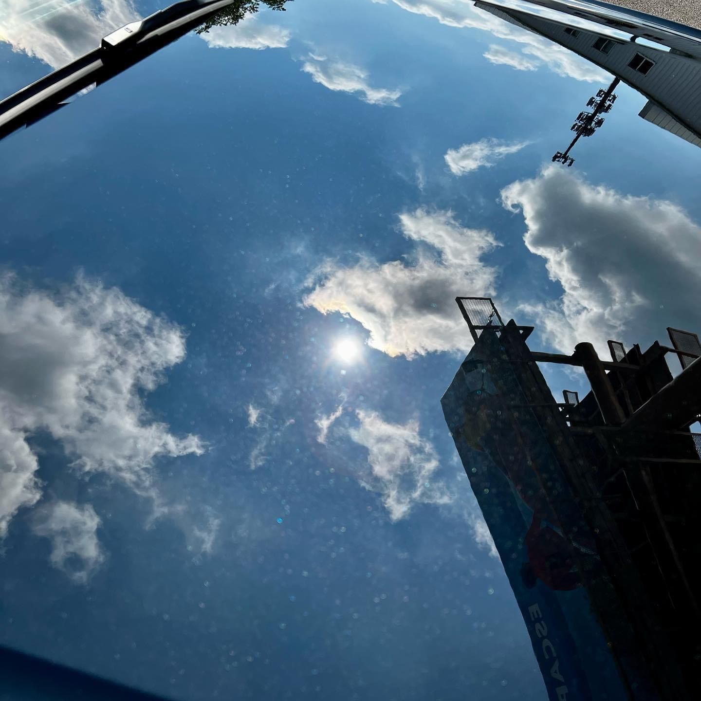 Blue sky with clouds reflected on a car windshield, with buildings and a crane at the edge of the frame