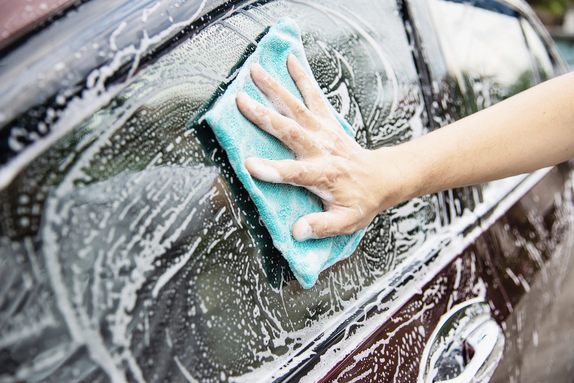 Hand washing a dark car window with a blue sponge and soap suds