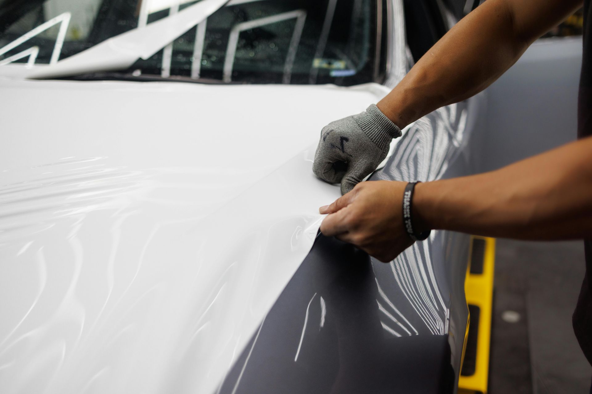 Hands applying white vinyl wrap to a car hood with a gray fender visible underneath