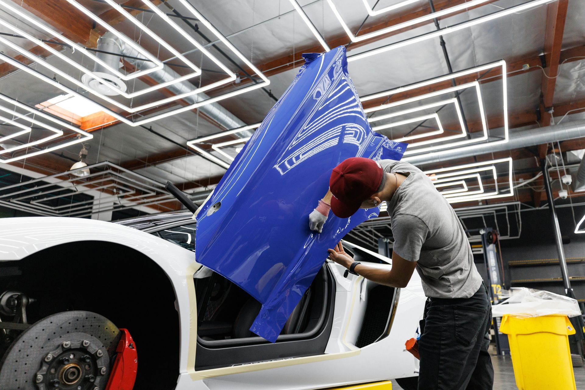 Worker polishing blue car panel in an auto shop with ceiling lights overhead