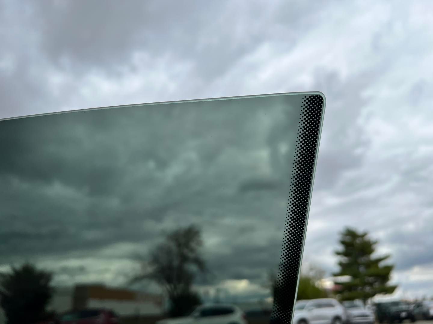 Dark cloudy sky seen through a car window, with parked cars and trees in the distance