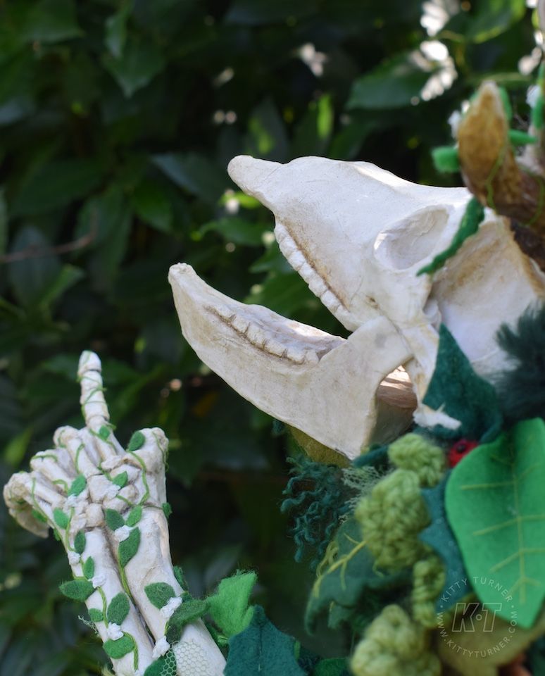 The paper mache skeleton puppet looking happy up at the sky and pointing. It has textile leaves covering it. 