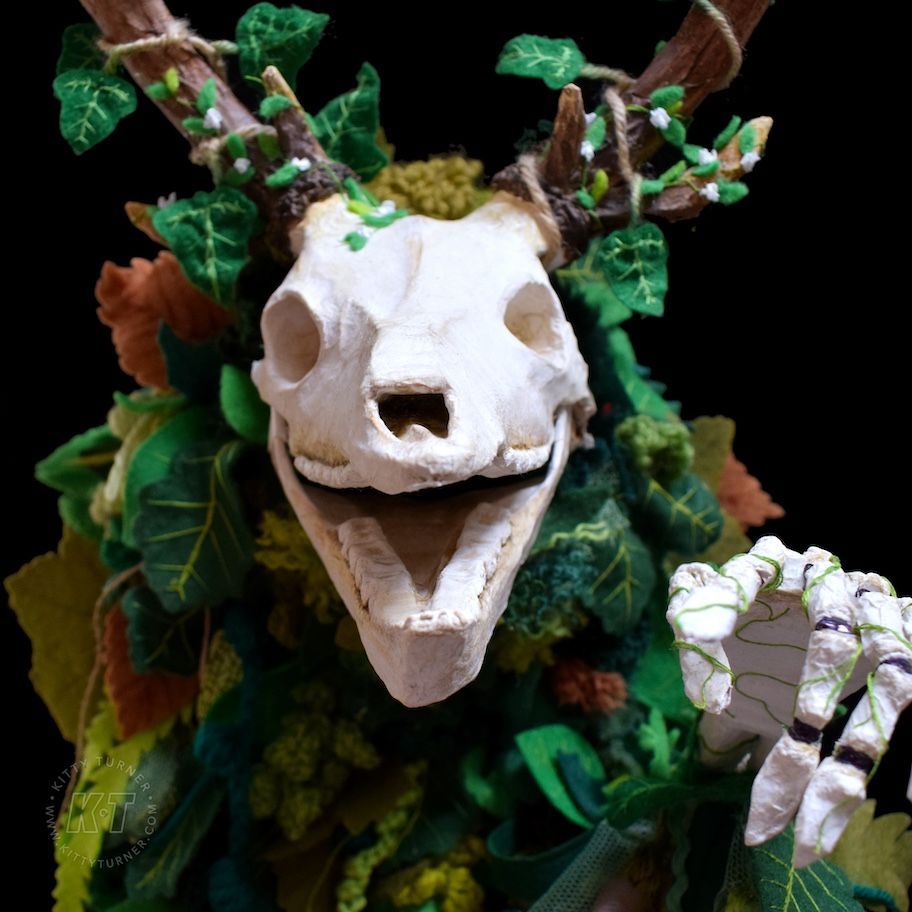 The paper mache skeleton puppet looking and pointing at the camera. It has handmade textile leaves covering it. 