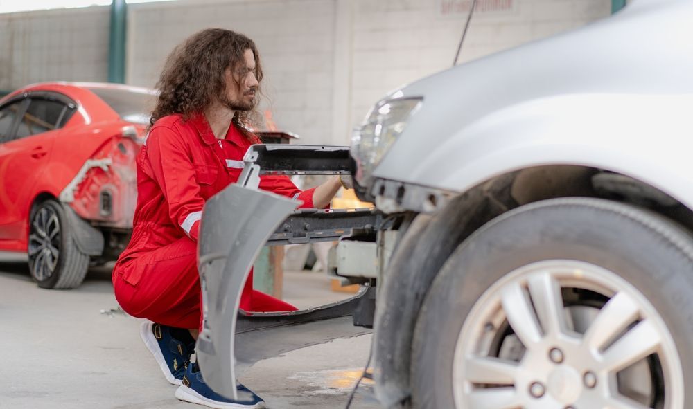 Mechanic in red coveralls working on a car in a repair shop, with a red car in the background.