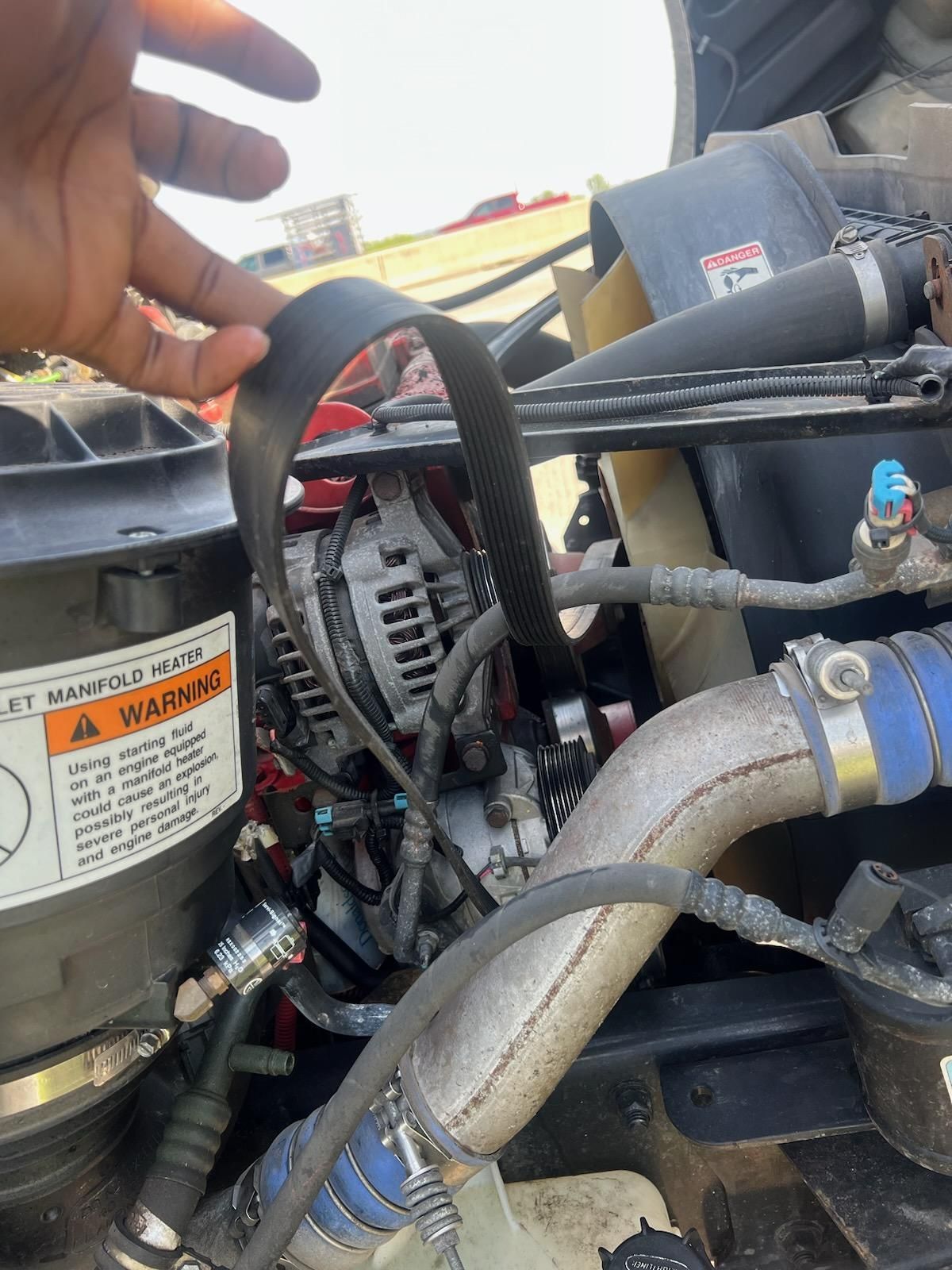 Close-up of a vehicle engine compartment with hoses, wiring, and a hand pointing inside.