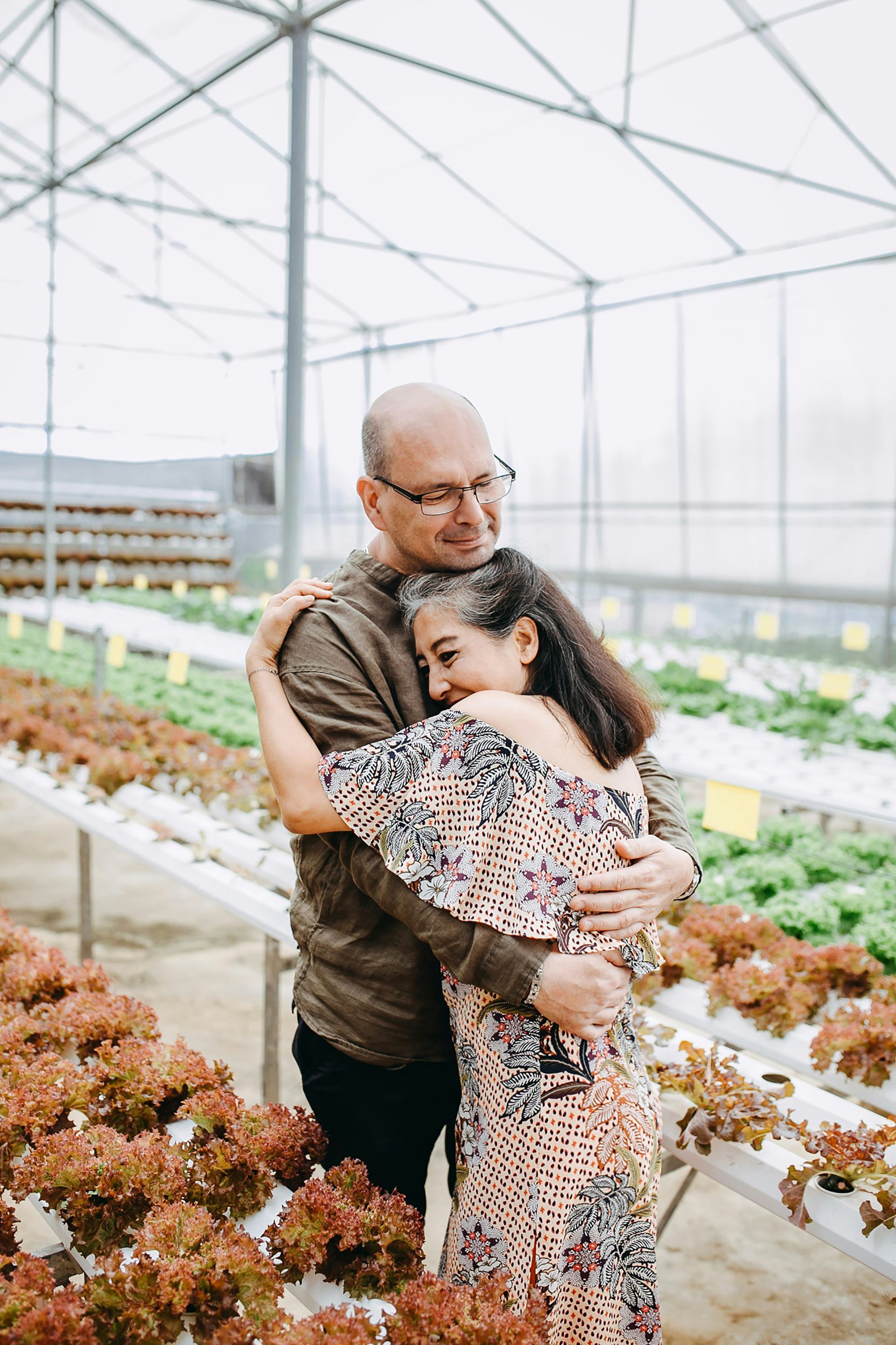 An elderly woman wearing a straw hat is standing in a greenhouse.