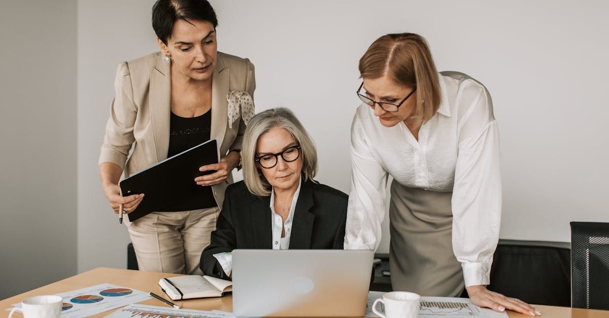 Three women are standing around a table looking at a laptop computer.