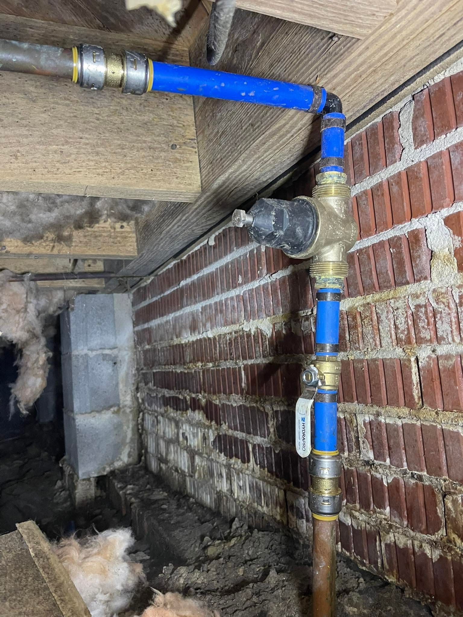 Blue and copper pipes with fittings running vertically and horizontally along a brick wall under a building.