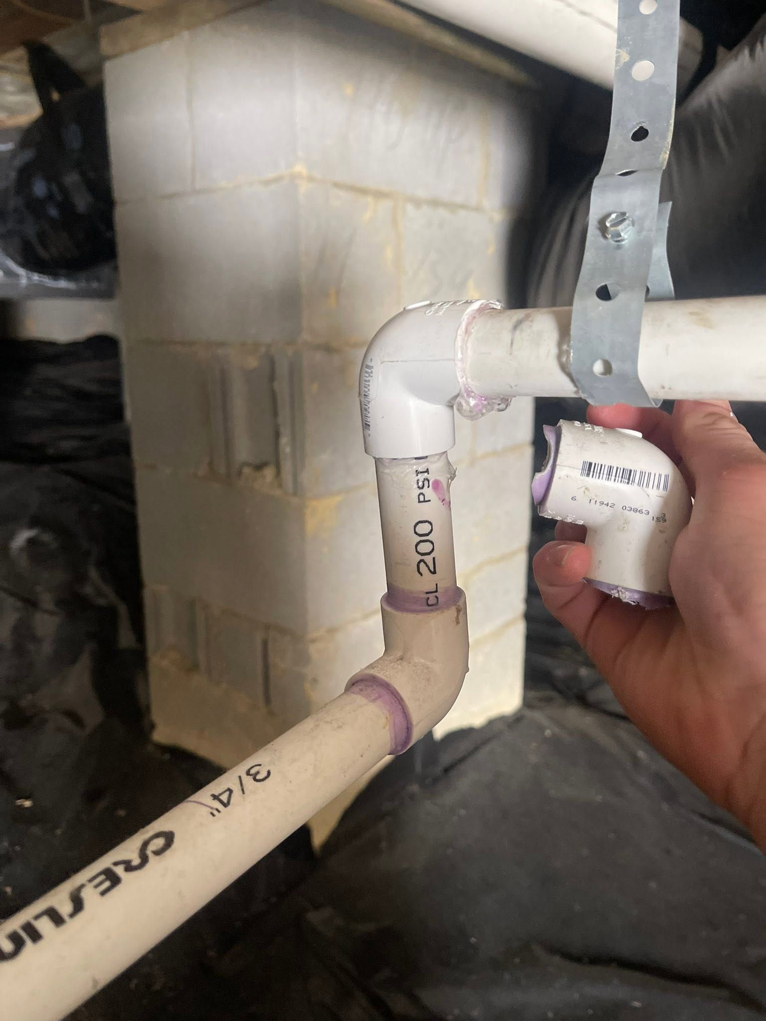 Hand installing a white PVC pipe elbow inside a utility space beside a gray HVAC duct or furnace.