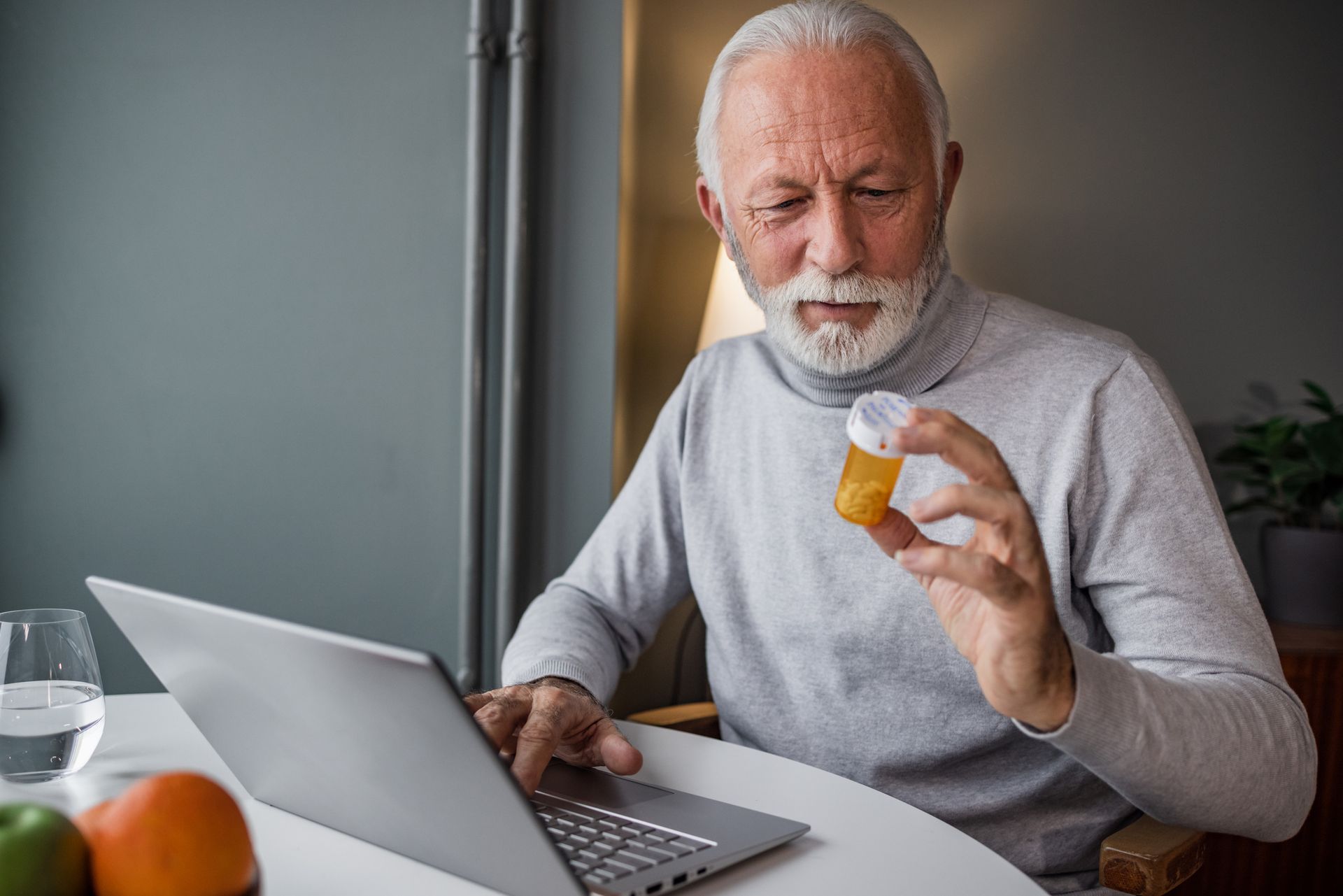 An elderly man is holding a bottle of pills while using a laptop computer.