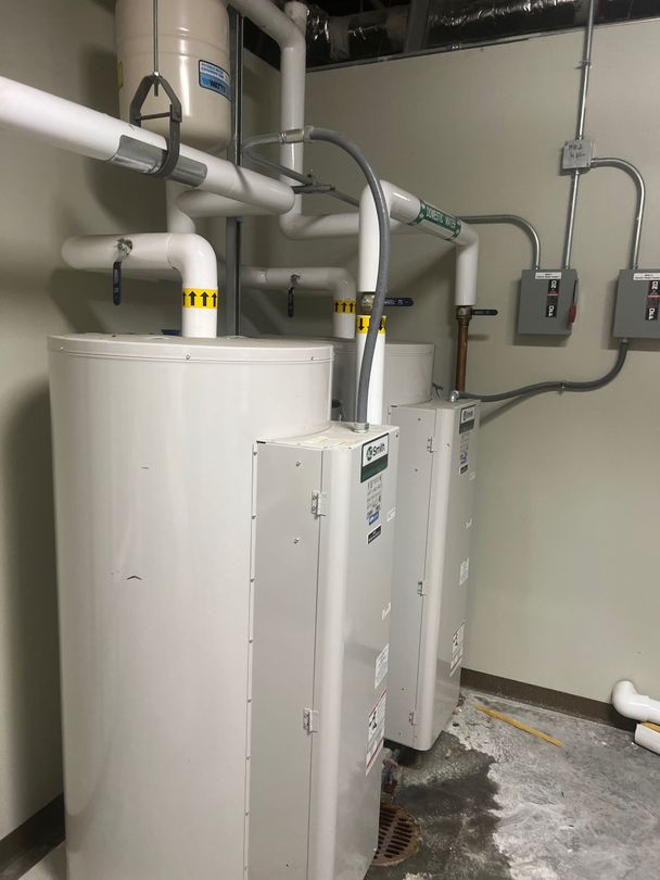 Three white water heaters and pipes in a utility room, with water pooled on the floor.