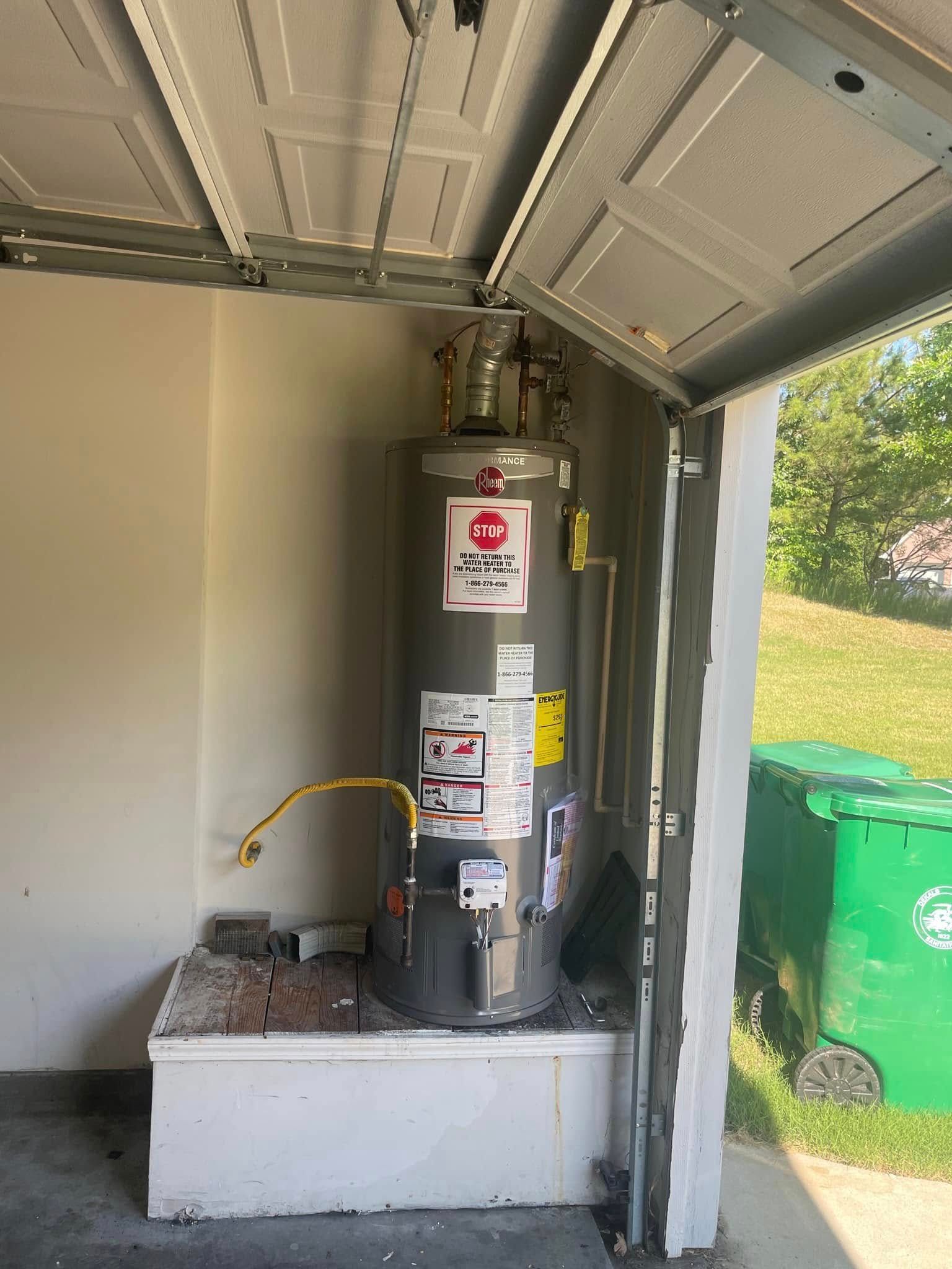 Water heater in a small outdoor utility shed, with a grassy yard and green trash bins outside.