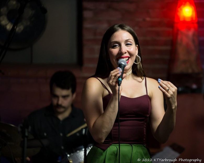 Singer performing onstage with microphone, smiling under warm red lights; musician blurred in background