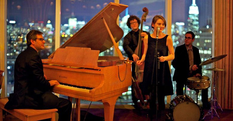 Jazz trio performing in front of city skyline, with a pianist at a grand piano and two singers beside a drummer.
