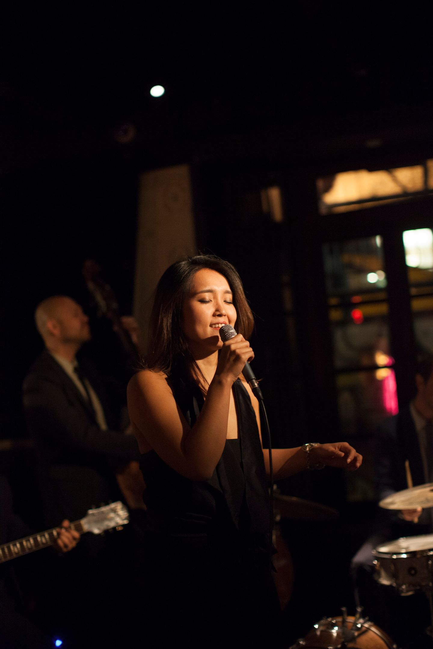 Singer performing on a dimly lit stage with a microphone, guitarist and drum set in the background