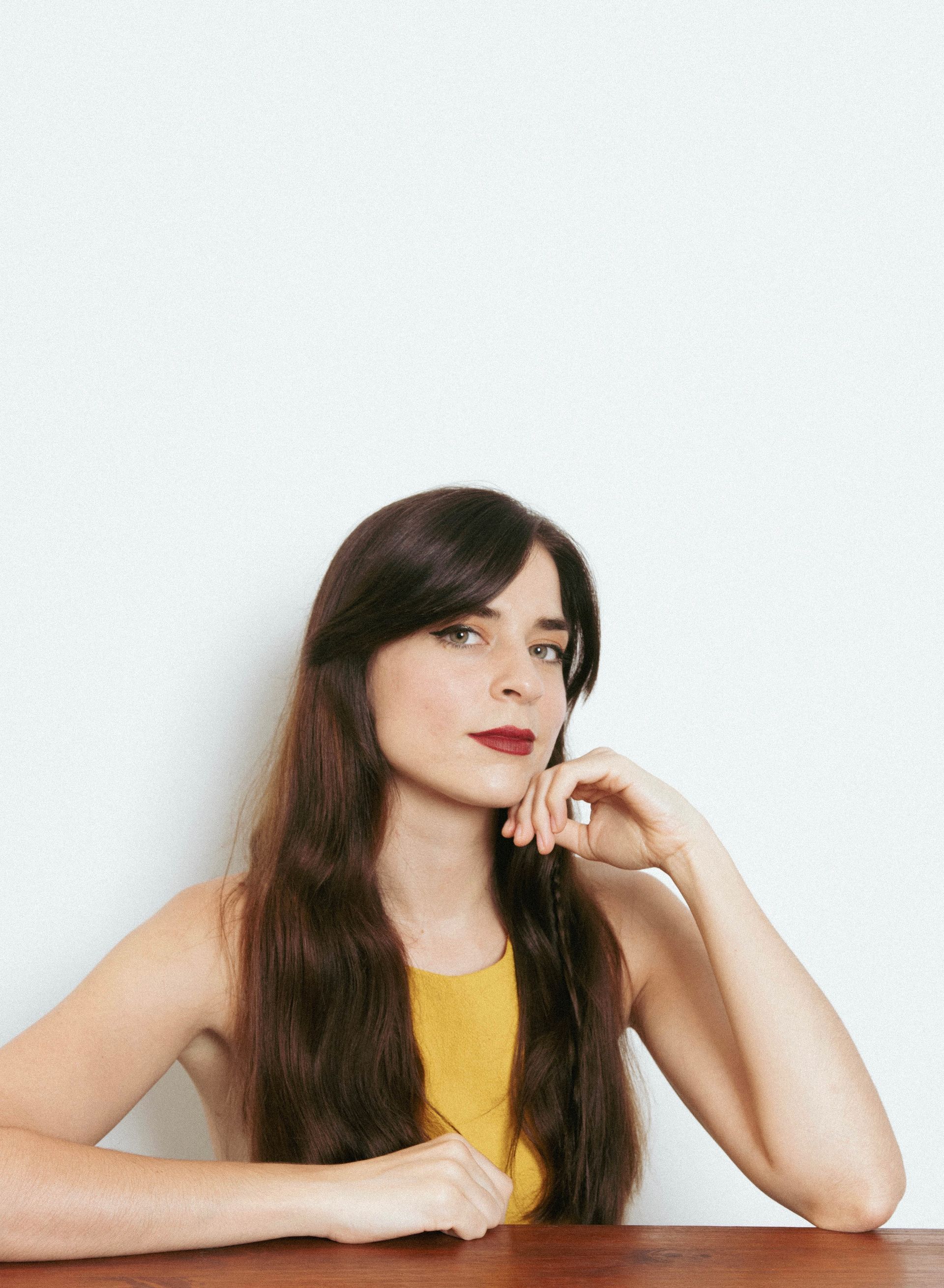 Woman with long dark hair in a yellow top resting her chin on her hand against a light background