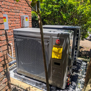 Outdoor HVAC unit beside a brick wall, with a young tree in front and greenery behind.