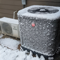 Snow-covered outdoor air conditioner units beside a house wall