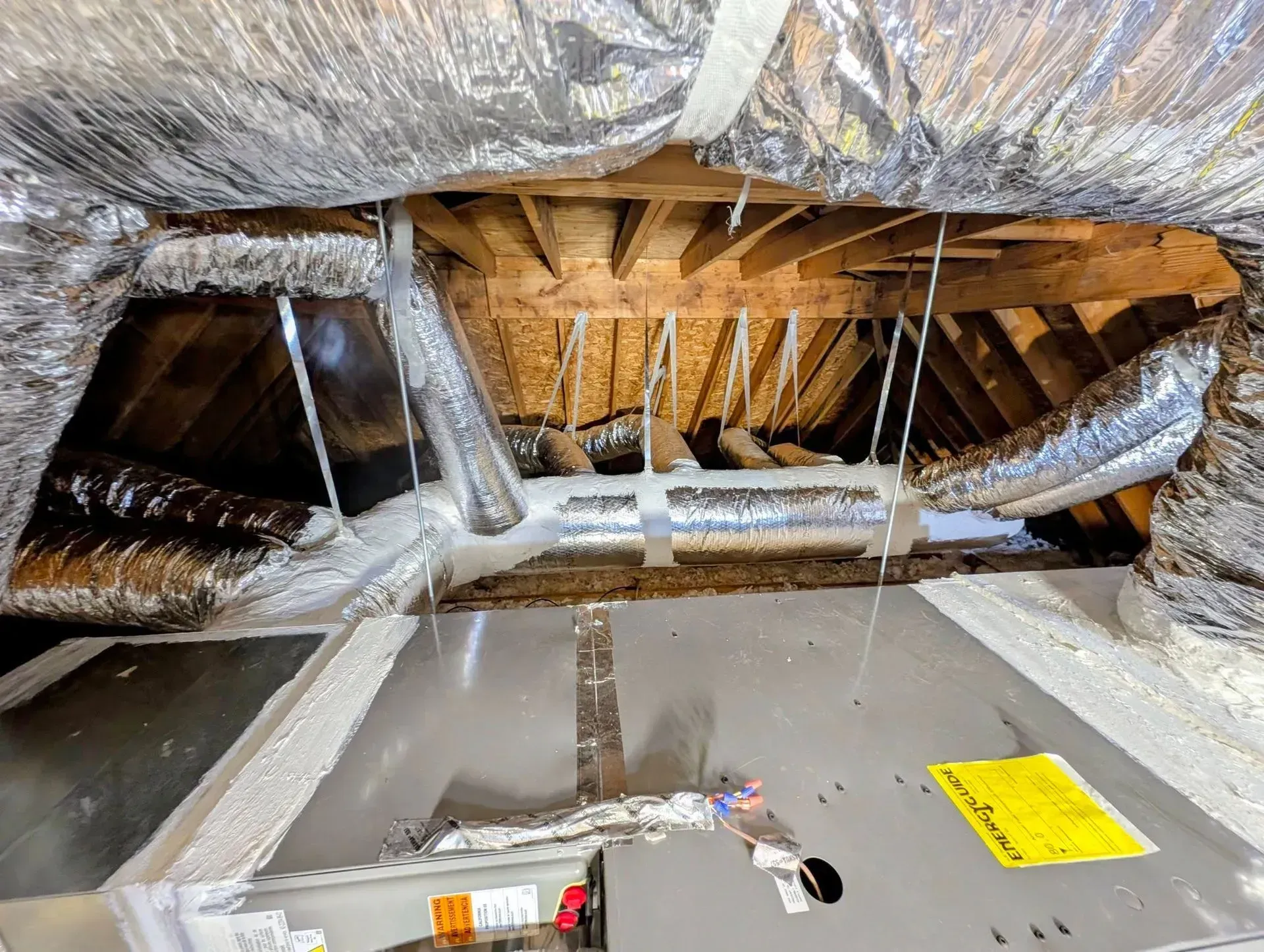 Insulated attic with exposed rafters, hanging ducts, and a yellow warning label on the floor.