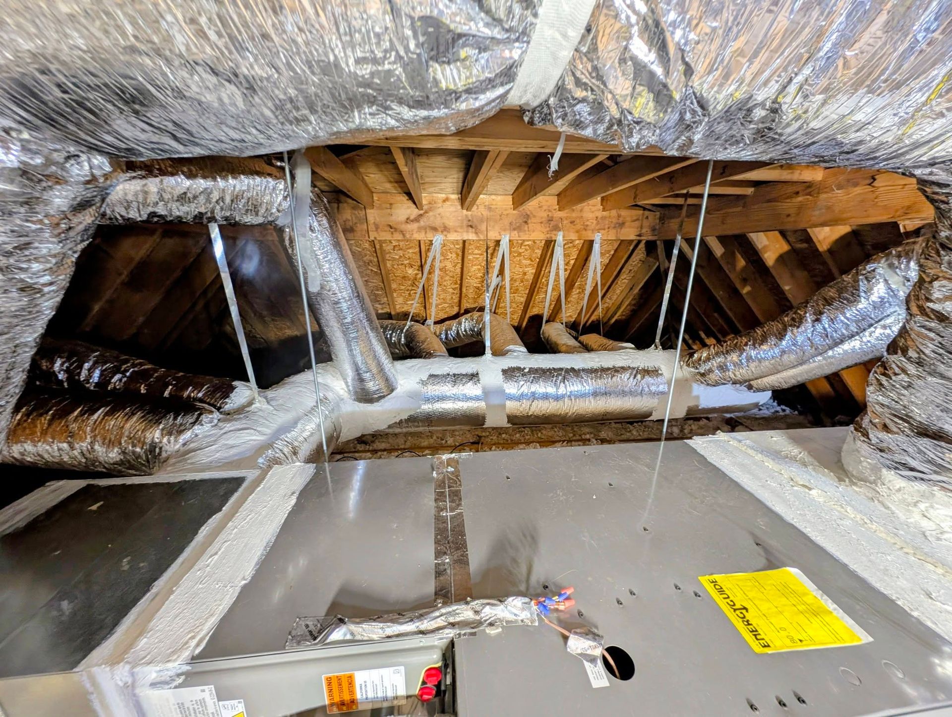 Insulated attic with exposed rafters, hanging ducts, and a yellow warning label on the floor.