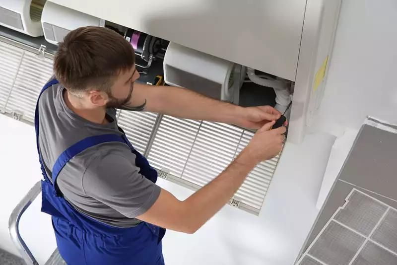 Technician in blue overalls adjusts a window blind mechanism on a wall-mounted unit.