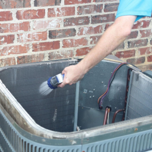 Person sprays water into an open HVAC unit beside a brick wall.