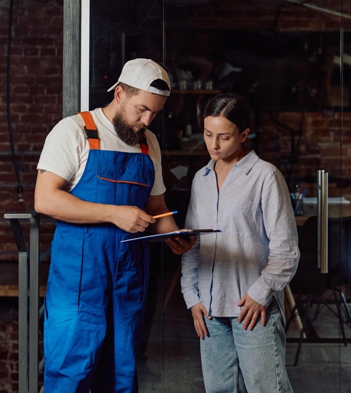 Two workers review a clipboard in a workshop, one pointing while the other listens.