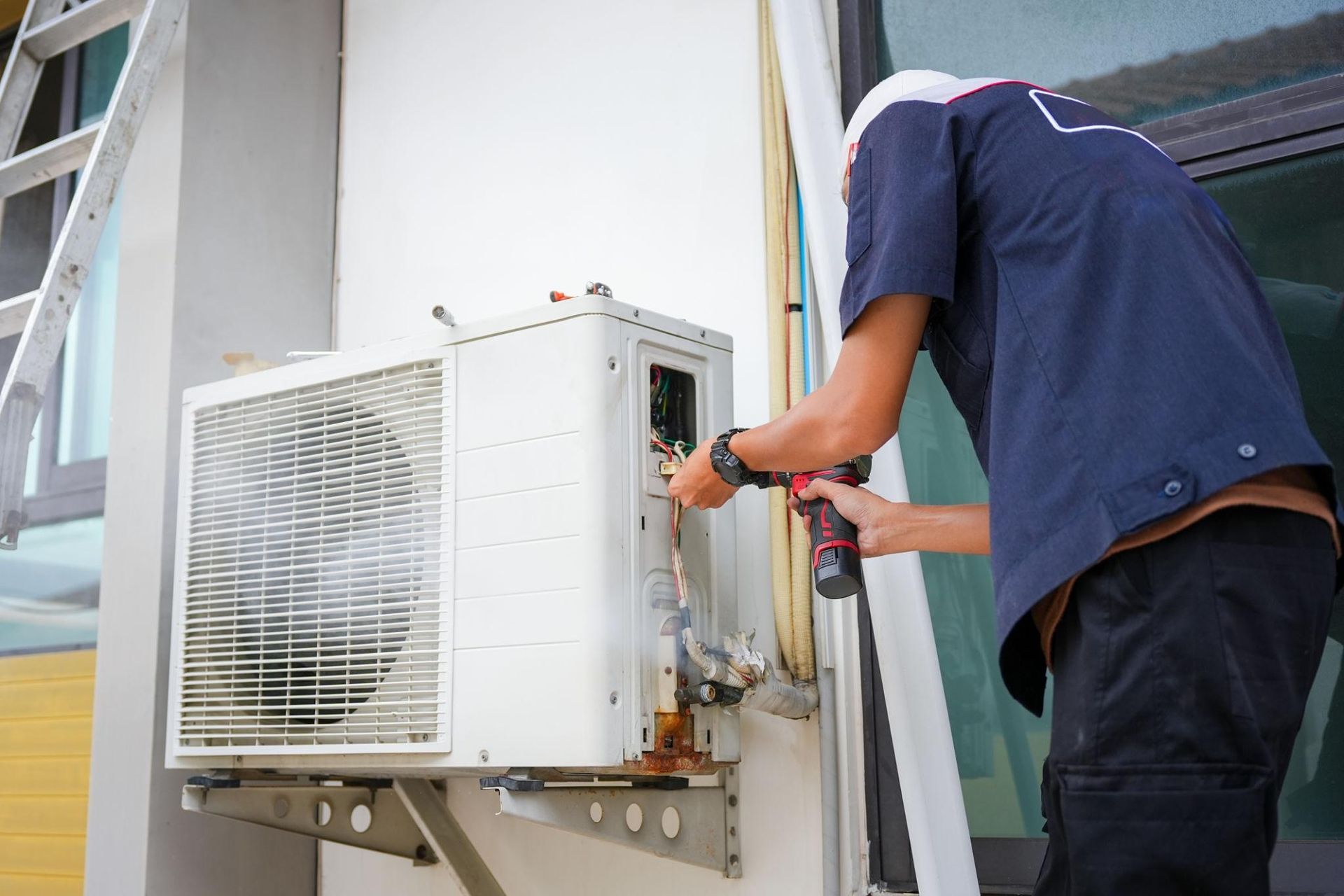 Technician repairing an outdoor air conditioner unit with tools near a building wall