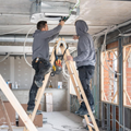 Two workers installing a ceiling light in an unfinished room with ladders and exposed walls