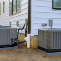 Two outdoor HVAC units beside a house with white siding and exposed utility pipes