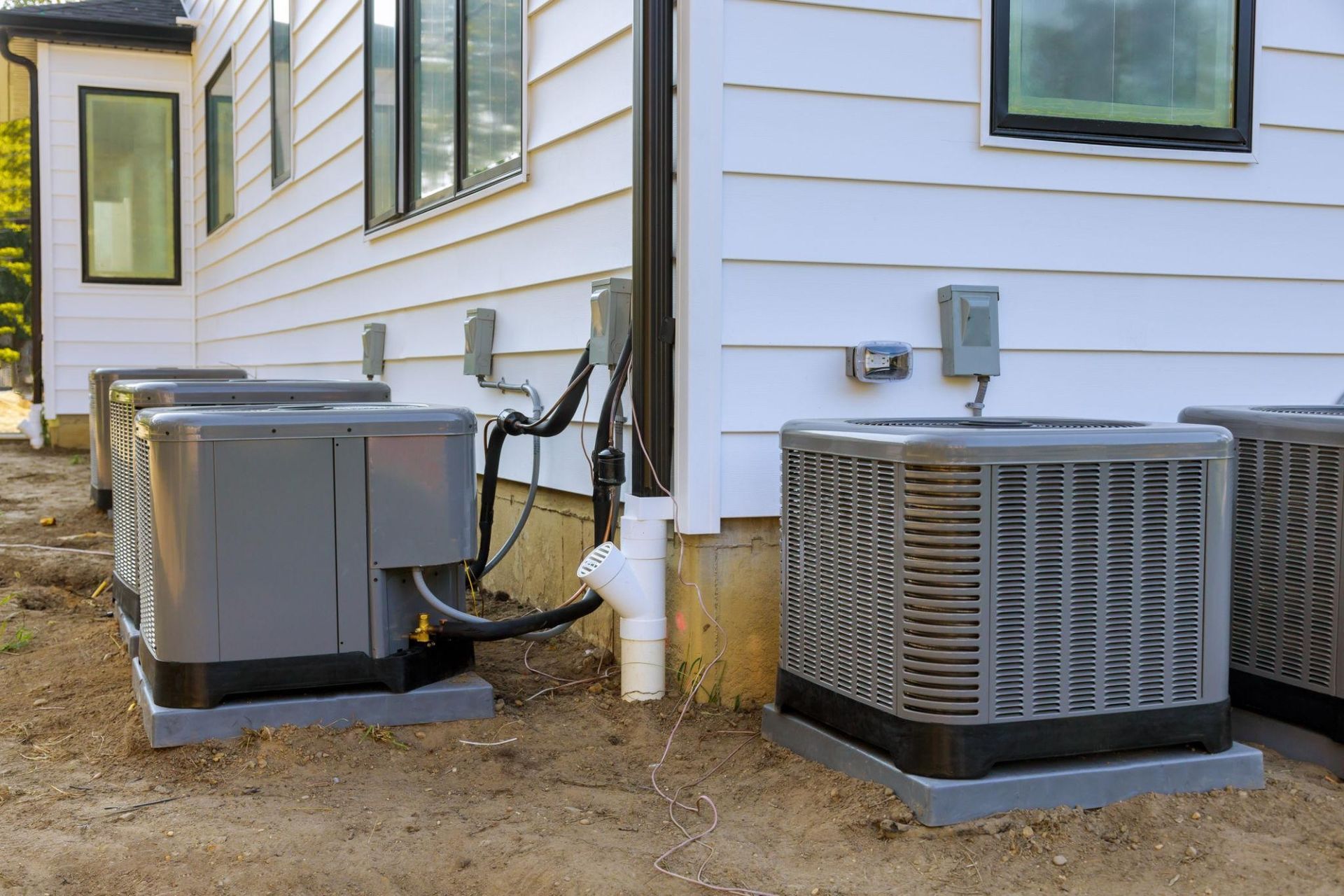 Two outdoor HVAC units beside a white house, with pipes and wiring visible.