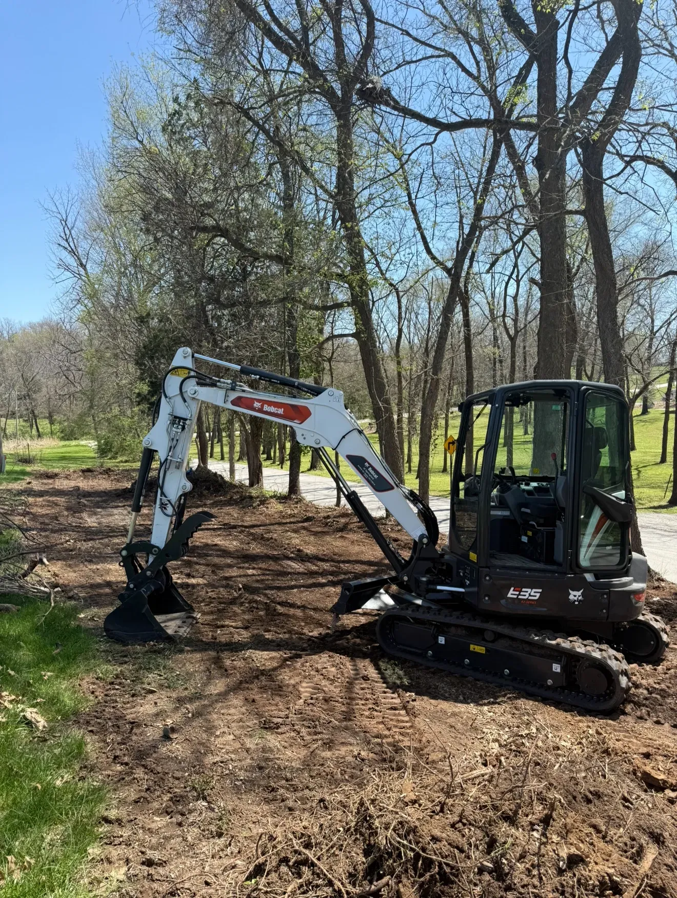 A white Bobcat mini-excavator with black tracks sits on a dirt clearing in a wooded area on a sunny day.