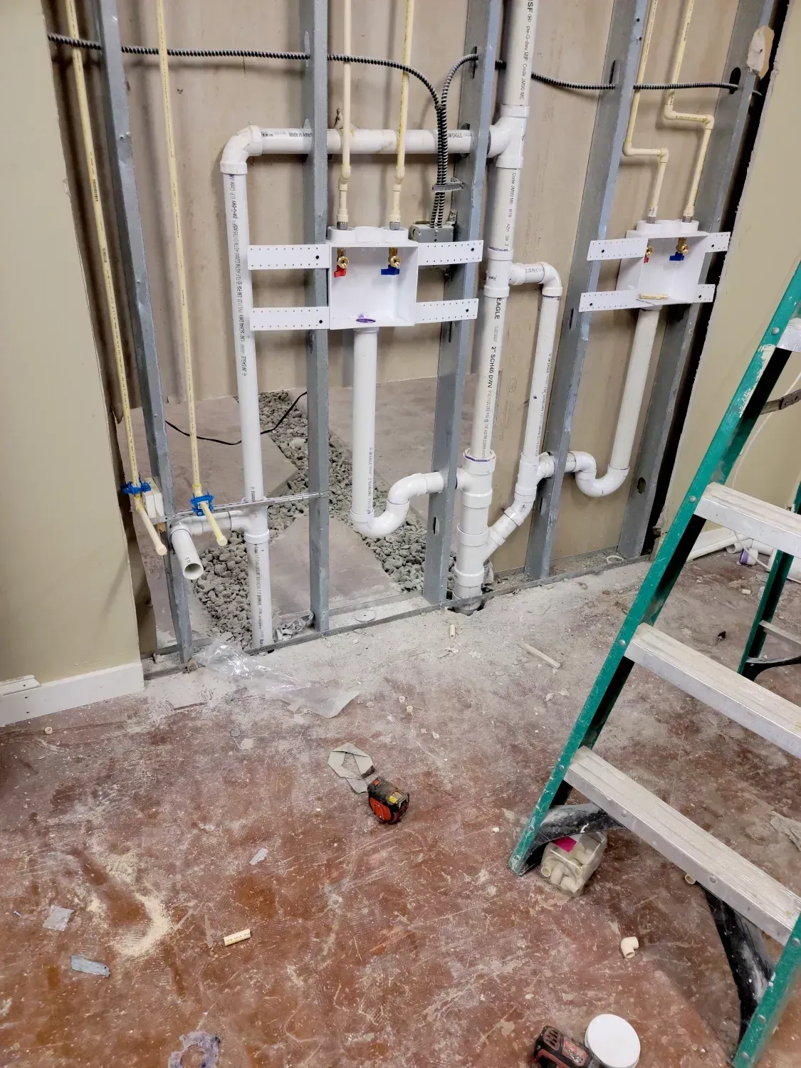 Plumbing rough-in with white PVC pipes, steel framing, and a green ladder inside a partially constructed wall.