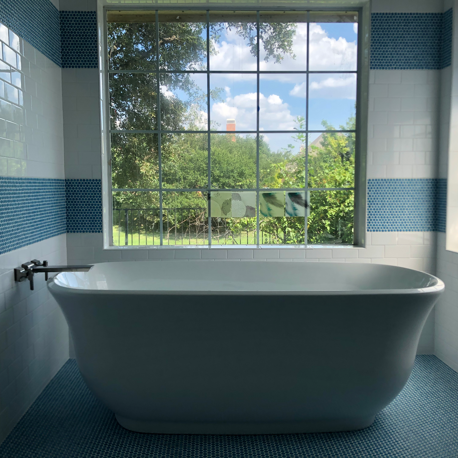 Clawfoot tub in bathroom with blue tiled walls, window overlooking green trees and blue sky.