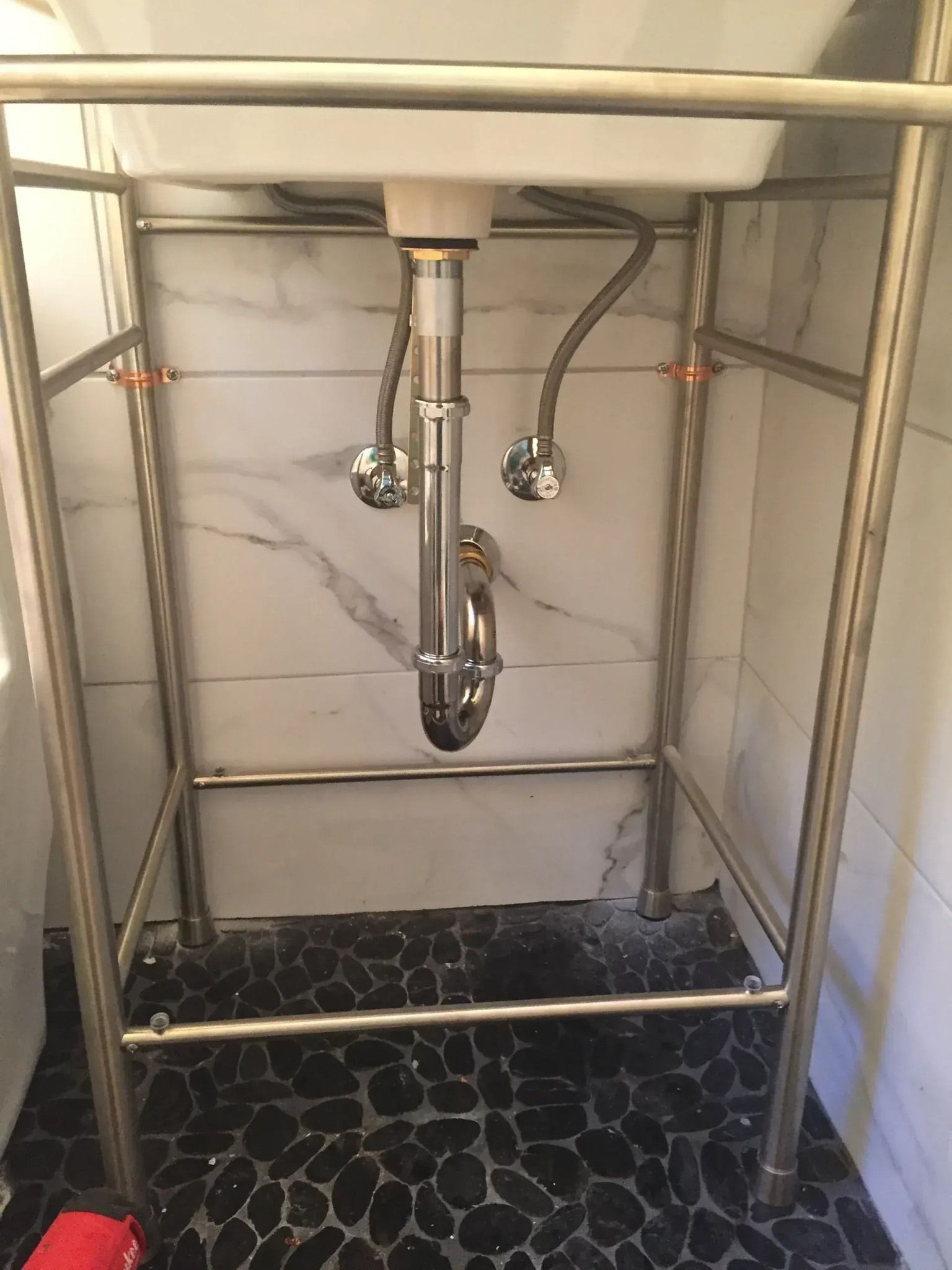 White sink on stainless steel stand; plumbing visible; against marble tile wall, black stone floor.