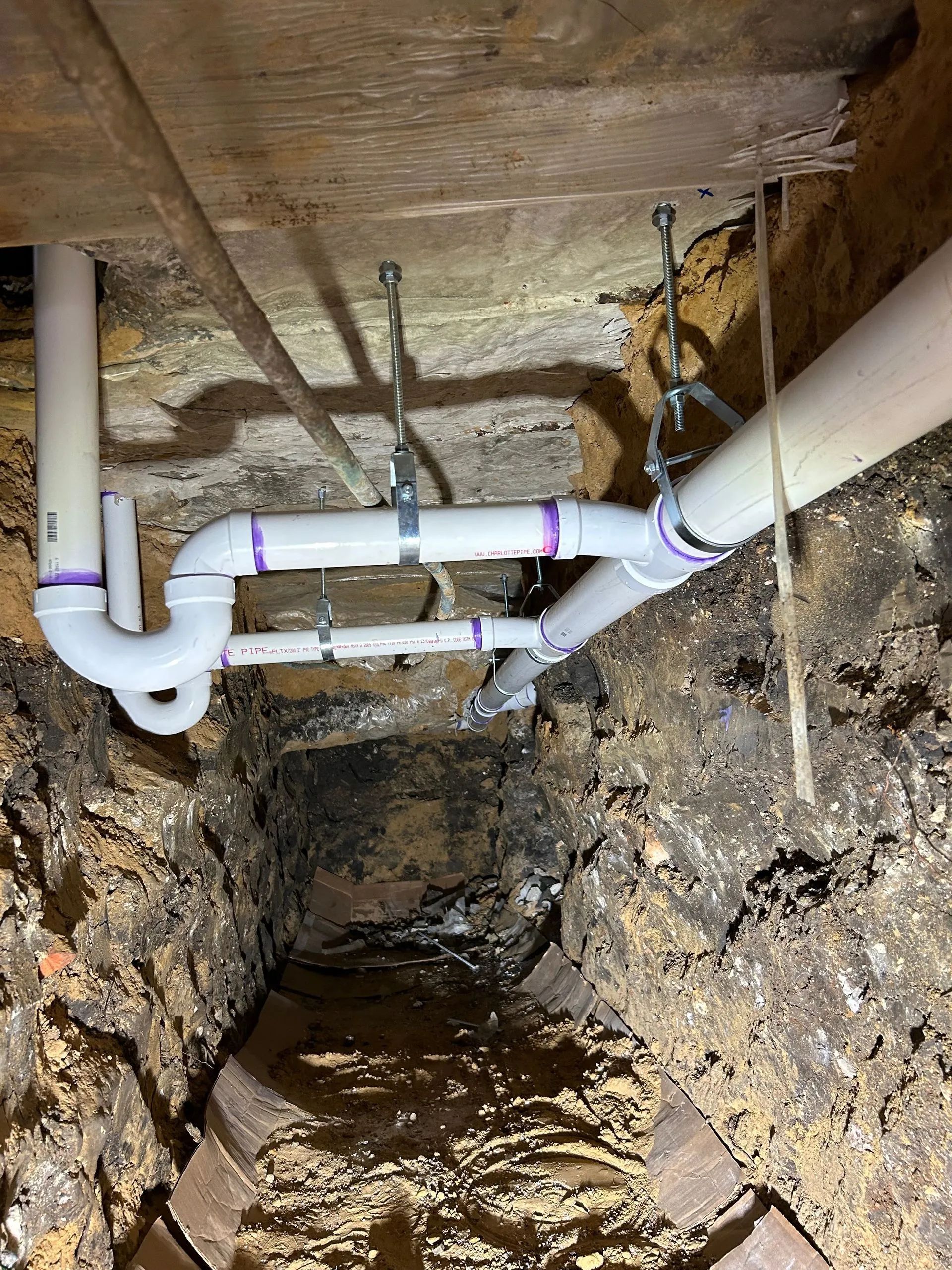 PVC pipes installed in a trench beneath a wooden floor, with exposed dirt walls and insulation above