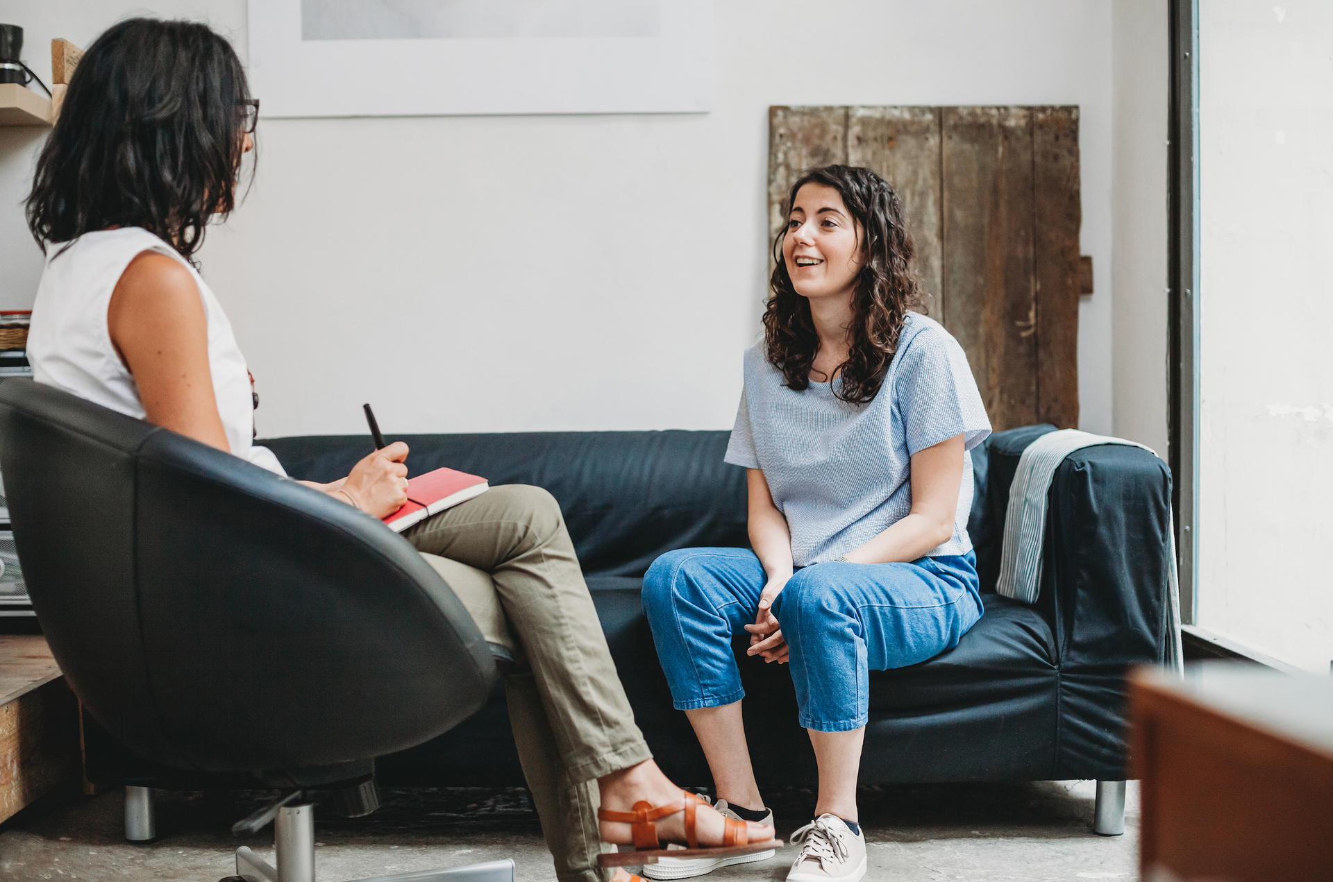 A woman is sitting on a couch talking to another woman.
